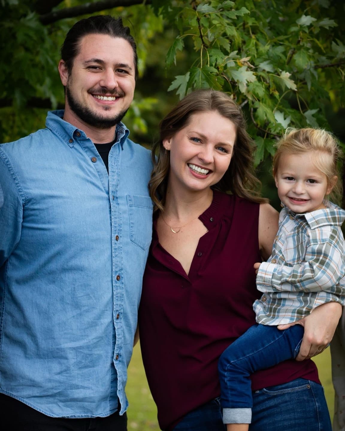 A young family of three smiling under the trees